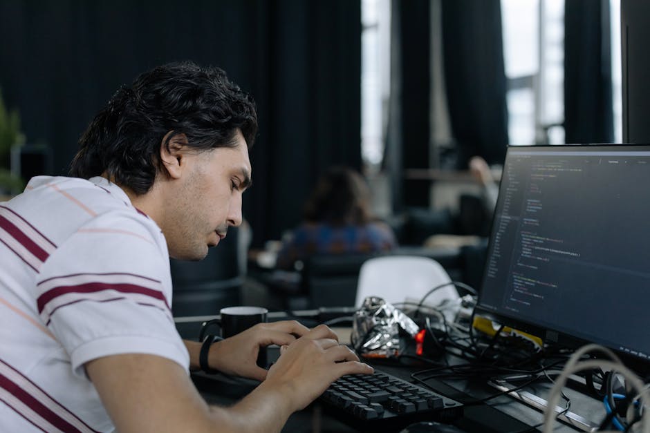 The image shows a young man with dark, curly hair working intently at a desk in an indoor office or classroom environment. His right hand is on a keyboard, while his left hand appears to be adjusting